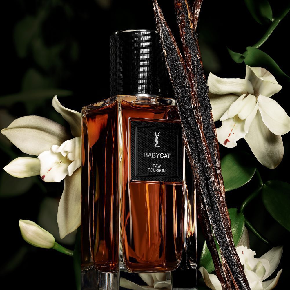 An amber-colored perfume bottle labeled "BABYCAT RAW BOURBON" by YSL, adorned with vanilla pods and white orchid flowers.