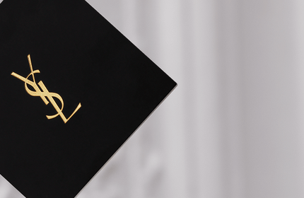 A close-up of luxury beauty products, including a compact and a liquid foundation, both featuring the YSL (Yves Saint Laurent) logo, against a stylish background.