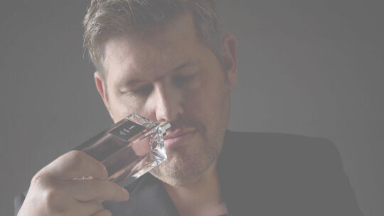 A man in a dark top holds a perfume bottle to his nose, deeply inhaling its scent.