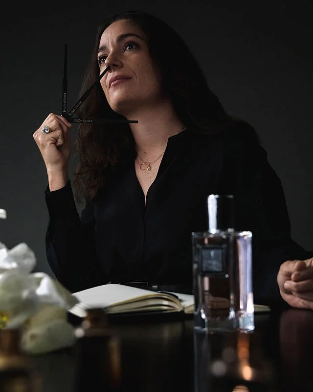 A woman in a black shirt holds a perfume blotter, looking up thoughtfully, with a notebook, perfume bottle, and flowers on the table.