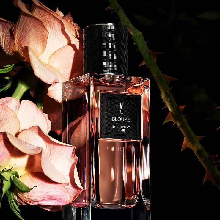 A rectangular perfume bottle labeled "BLOUSE IMPERTINENT ROSE" by YSL, surrounded by soft pink roses and thorny rose stems.