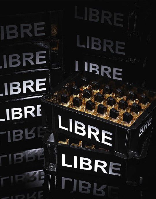 A composition of black crates, similar to those used for transporting bottles, stacked and filled with YSL Libre perfume bottles. The crates bear the inscription "LIBRE" in large, luminous white letters. In the foreground, one crate is filled with multiple "Libre" perfume bottles that reflect light, creating golden glints. The entire scene is bathed in dim light, accentuating the chic aspect and the product's staging.