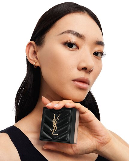 A woman holds a black, square Yves Saint Laurent compact near her face.