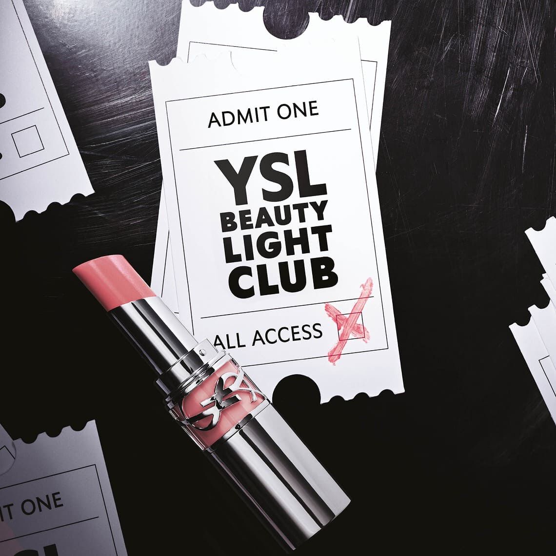 A light pink lipstick with a glossy texture rests on two slightly overlapping white event-style tickets. The top ticket features "ADMIT ONE" at the top, and "YSL BEAUTY LIGHT CLUB" in large black letters in the center, with "ALL ACCESS" below. A red cross is scribbled on the bottom right corner of the ticket, as if validating it. The background is dark, suggesting a textured surface, and torn ticket edges are visible on the sides, hinting at several other scattered tickets. The overall impression is one of an exclusive invitation to a glamorous event.