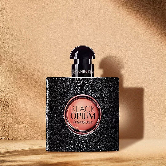 A Black Opium perfume bottle with a glittery black finish and a round pink label displaying "BLACK OPIUM YVES SAINT LAURENT" in the center. The bottle has a dark cap and sits on a wooden surface against a light beige background with shadows.