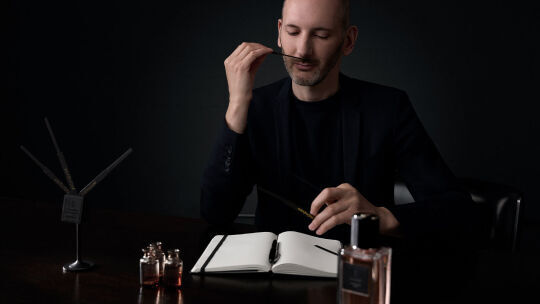 A perfumer in a dark suit attentively smells a perfume blotter in a dimly lit setting.