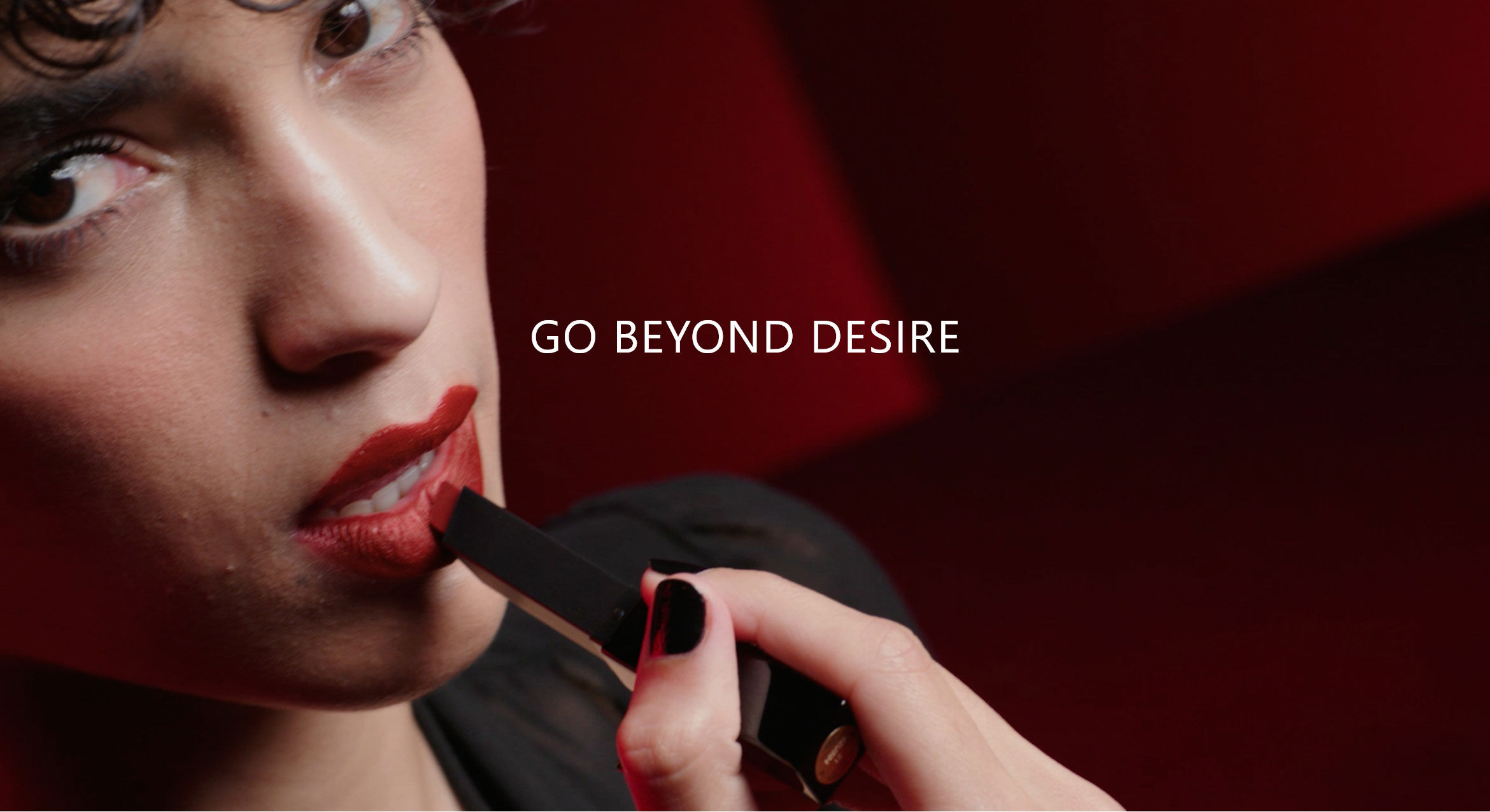 A close-up of a person applying red lipstick, with the phrase "GO BEYOND DESIRE" prominently displayed above. The background features rich shades of red, creating a bold and dramatic atmosphere.