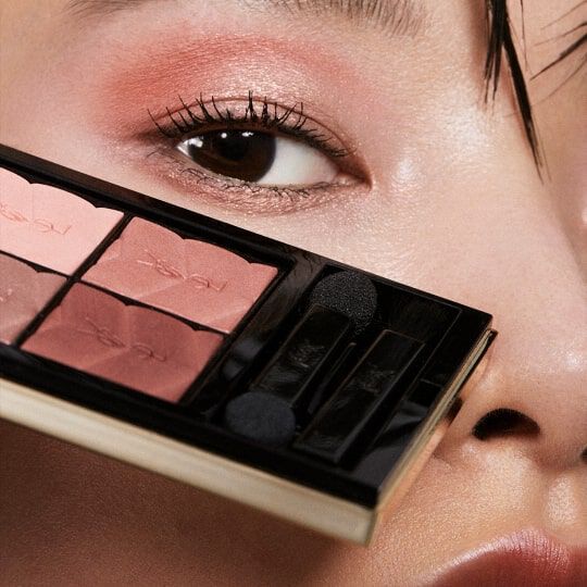 Close-up of an eye adorned with soft pink eyeshadow, holding a makeup palette featuring shades of pink and peach near the face.