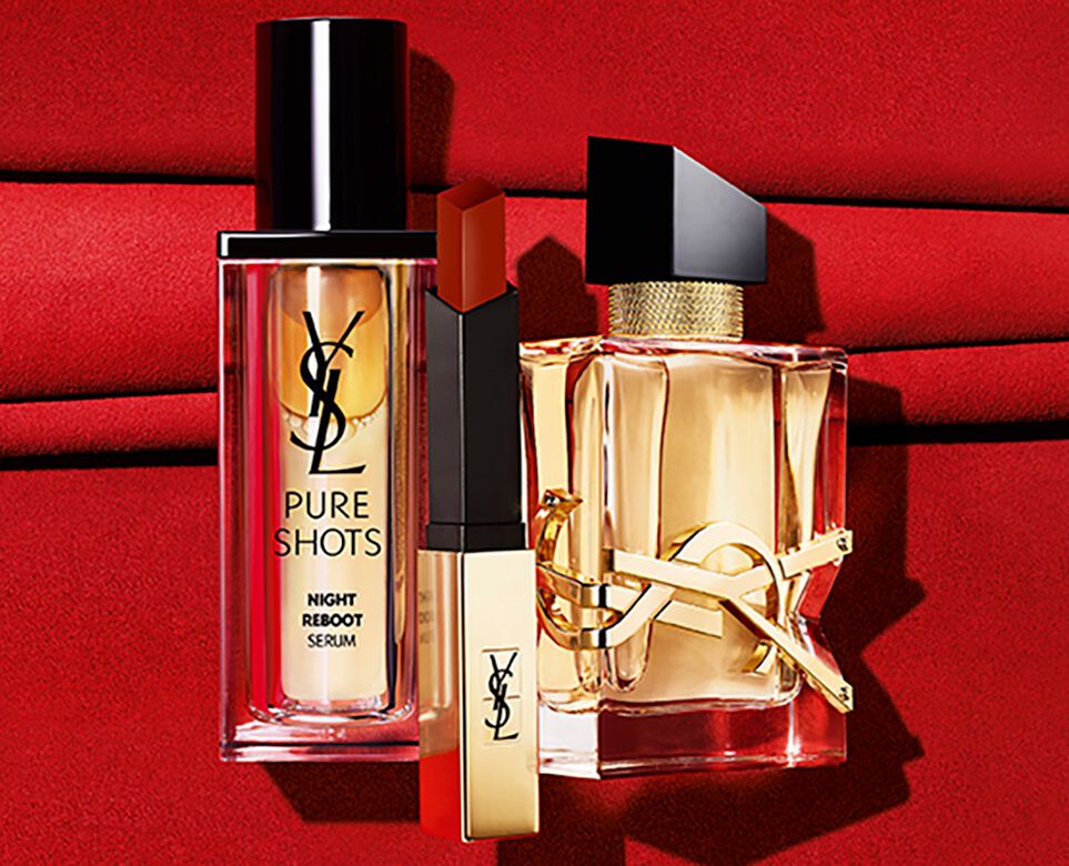 A close-up of two skincare bottles and a lipstick on a rich red fabric backdrop. The products are from the YSL brand, featuring sleek, modern packaging.