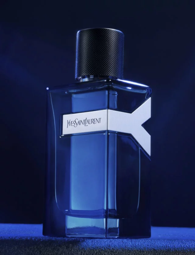 A close-up of a blue Yves Saint Laurent perfume bottle against a dark, smoky background. The bottle has a sleek design with a prominent white label featuring the brand name.