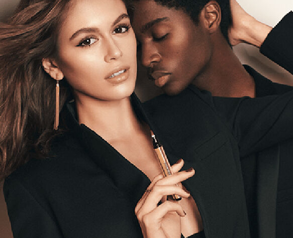 A close-up shot of a woman and a man posing together against a neutral background, showcasing stylish outfits. The woman has long hair and is holding a makeup brush, while the man is leaning in closely with a soft expression.