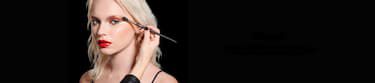 A person applies makeup to a model's cheek, who has long blonde hair and striking red lips, against a dark background.