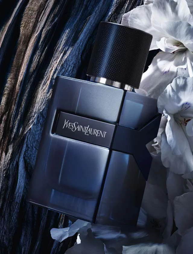A dark, stylish perfume bottle sits atop a textured wooden surface, surrounded by white flowers. The bottle has a sleek design with a metallic cap and features the brand name "Yves Saint Laurent" prominently displayed.