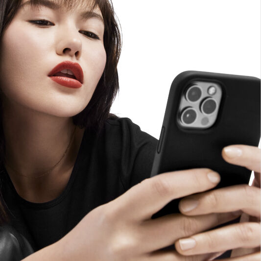 A close-up of a woman with dark hair and bold red lipstick, holding a smartphone and looking at the screen with a thoughtful expression. She is wearing a black shirt.