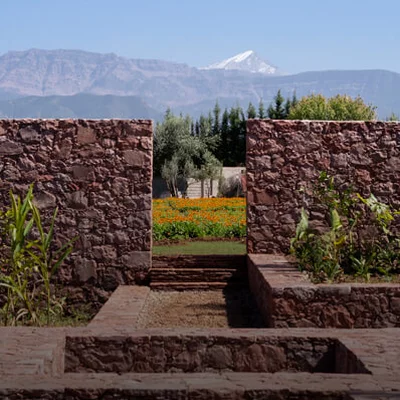 A view of a stone wall with an opening, revealing a colorful flower garden and snow-capped mountains in the background under a clear blue sky.