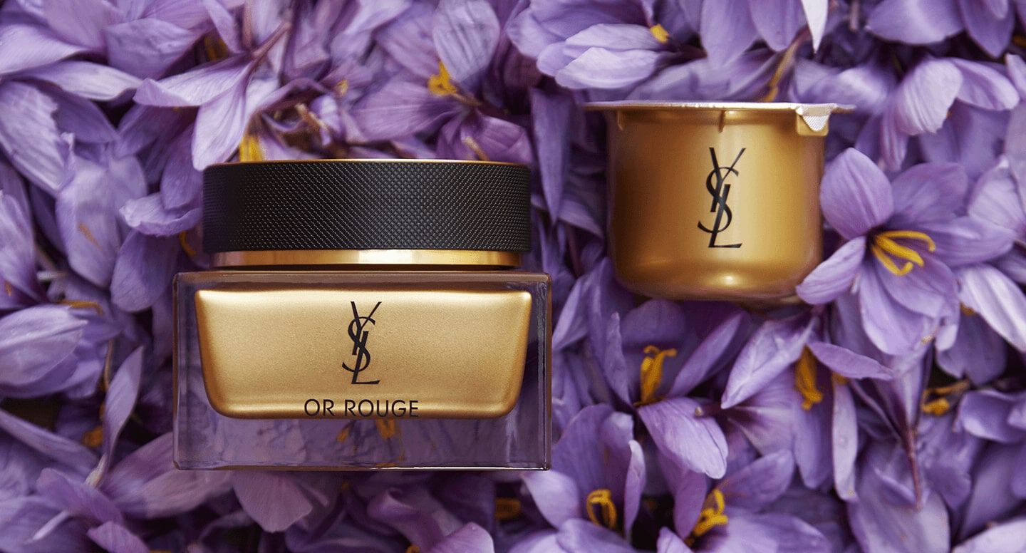A YSL Or Rouge skincare jar and cap are displayed elegantly on a bed of purple flower petals.