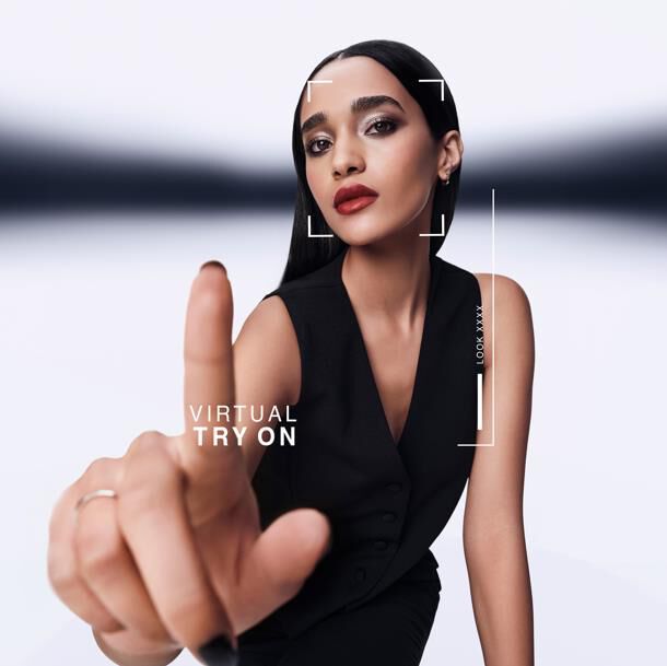 A woman points forward, interacting with a virtual try-on screen. A graphic overlay shows "VIRTUAL TRY ON" and "LOOK XXXX" around her face. She wears a black vest.​