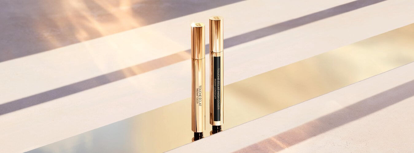 Two elegant gold and black makeup pens are positioned upright on a reflective surface, with soft light and a warm background creating an elegant ambiance.