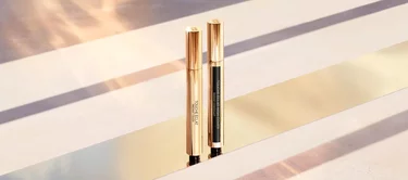 Two elegant gold and black makeup pens are positioned upright on a reflective surface, with soft light and a warm background creating an elegant ambiance.