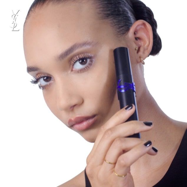 A young woman with dark hair is holding a sleek, black cosmetic product near her face, showcasing a bold makeup look with subtle eye highlights and glossy lips.
