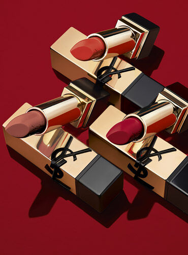 Three luxurious lipstick tubes by Yves Saint Laurent, featuring gold packaging and a prominent YSL logo, set against a rich red background.