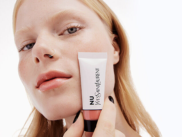 A model holding a tube of nude-colored makeup, showcasing a natural look with soft lighting and a neutral background.
