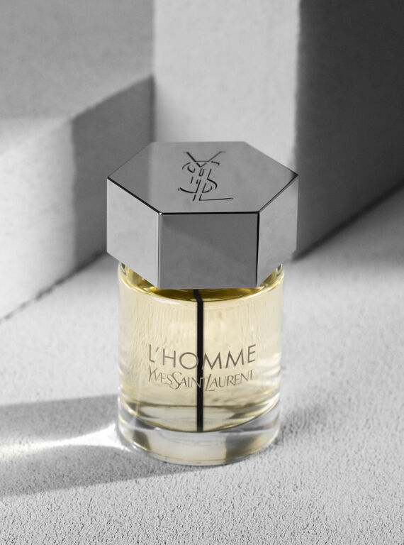 A close-up of an elegant glass perfume bottle with a hexagonal silver cap, labeled "L'Homme" by Yves Saint Laurent, sitting on a textured surface with soft lighting.