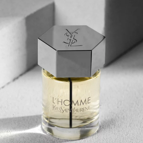 A close-up of an elegant glass perfume bottle with a hexagonal silver cap, labeled "L'Homme" by Yves Saint Laurent, sitting on a textured surface with soft lighting.