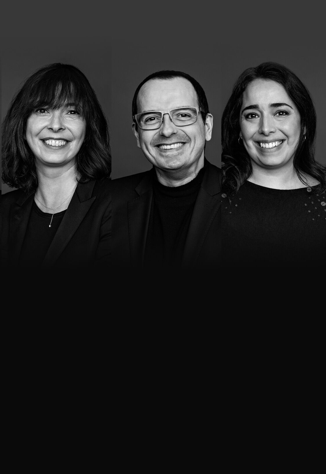 A black and white portrait featuring four individuals, each smiling, arranged in two groups of two. They are dressed in professional attire against a dark background.