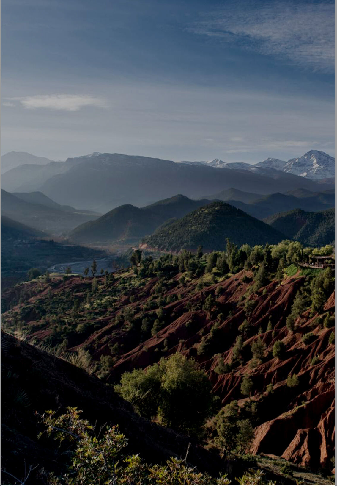 A panoramic view of a mountainous landscape with a fortress-like building in the foreground, surrounded by lush green hills and snow-capped peaks in the distance under a clear sky.