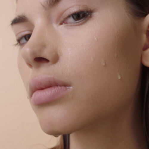 A close-up of a person's face with a natural look, showcasing dewy skin with beads of water running down the cheek.