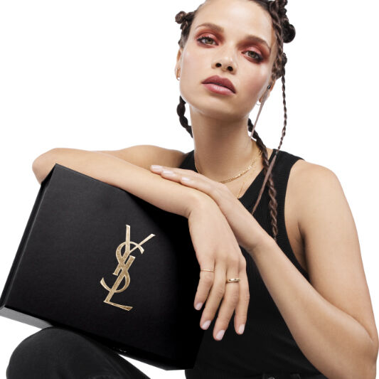 A model with braided hair poses elegantly while holding a black Yves Saint Laurent box, wearing a black tank top and bold makeup.