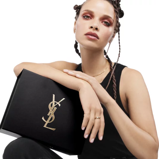 A model with braided hair poses elegantly while holding a black Yves Saint Laurent box, wearing a black tank top and bold makeup.