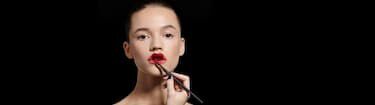 A model with a smooth, powdered face is having bright red lipstick applied to her lips by a makeup artist against a dark background.