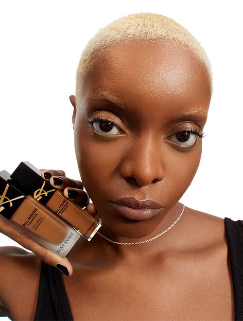 A close-up of a model holding a tube of YSL All Hours Concealer, showcasing her natural makeup look and clear skin.