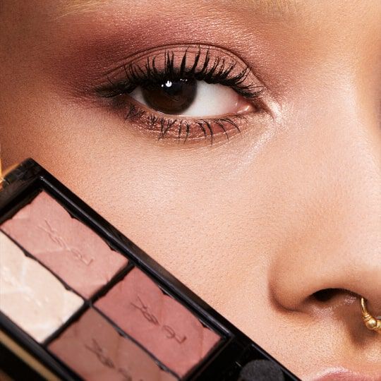Close-up of a person's eye with makeup, featuring long eyelashes and a subtle eyeshadow palette partially visible in the foreground.