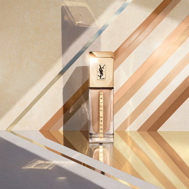 A stylish bottle of foundation set against a minimalist background with geometric stripes in soft beige and gold tones.