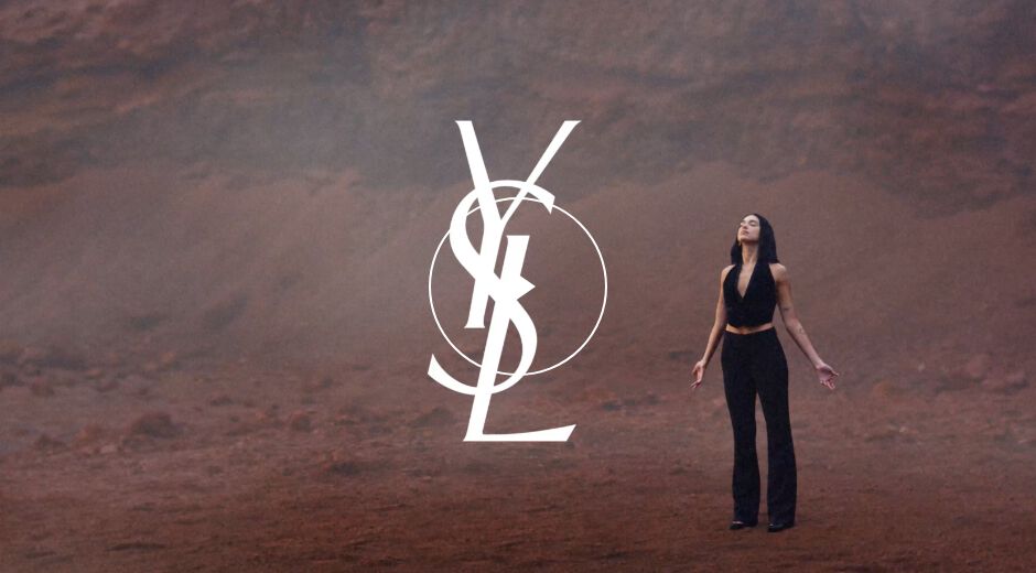 A woman in a black outfit stands with her arms raised, gazing upward against a dramatic, muted landscape with an oversized YSL logo overlay.