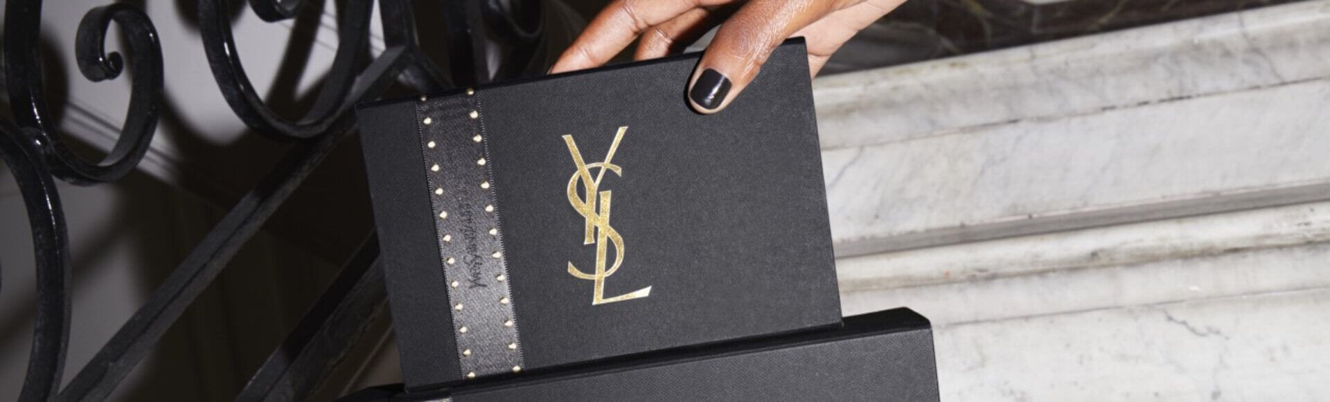 A hand holds a sleek, black gift box adorned with a gold YSL logo and a studded black ribbon, positioned against a marble background.