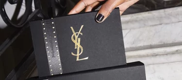 A hand holds a sleek, black gift box adorned with a gold YSL logo and a studded black ribbon, positioned against a marble background.
