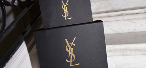 A hand with dark nail polish is placing a black box with a gold "YSL" logo on a marble staircase.