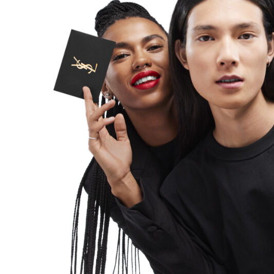 A confident young woman with bold red lips and braided hair holds a black card while posing alongside a young man with long, straight hair, both dressed in black attire against a neutral background.