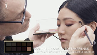 A makeup artist applies eyeshadow to a woman's eyelid using a brush, while she looks intently at the mirror. A palette of eyeshadow shades is visible in the foreground.