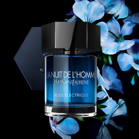 A bottle of Yves Saint Laurent's "La Nuit de l'Homme Bleu Electrique" perfume, featuring a sleek, dark cap and a blue glass design, surrounded by blue flowers on a black background.