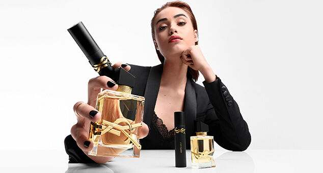 A person in a black blazer poses holding a large perfume bottle and smaller bottle, showcasing a stylish and sophisticated look.