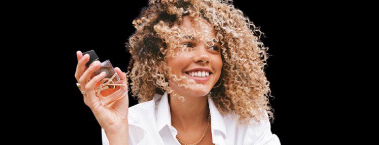 A woman with curly hair and a bright smile holds a small object in her hand, dressed in a white shirt against a dark background.