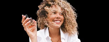 A woman with curly hair and a bright smile holds a small object in her hand, dressed in a white shirt against a dark background.