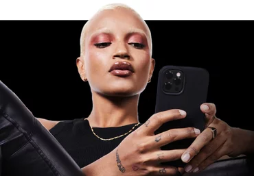 A person with short blonde hair and bold makeup is sitting and holding a smartphone, looking intently at the screen. They are wearing a black top and appear confident and engaged.
