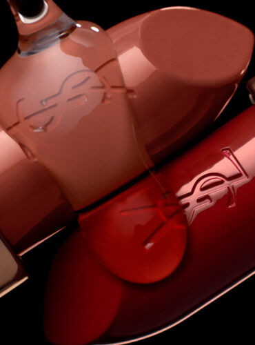 A close-up of several elegant lipstick tubes in varying shades of nude and red, featuring the YSL logo prominently on their surfaces, against a dark background.