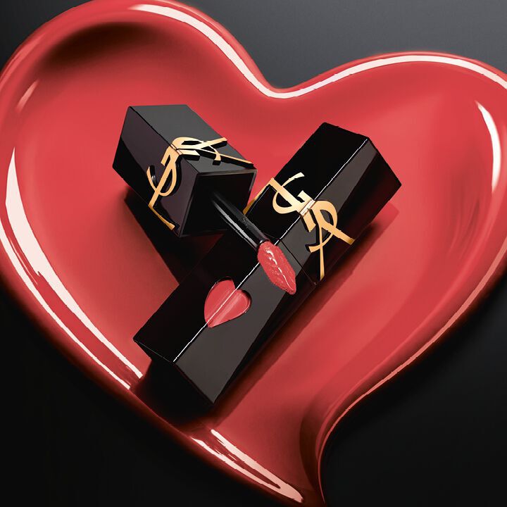 Two matte black YSL lip stain bottles with pink heart cutouts and gold logos, displayed inside a matching sculpted matte pink heart on a black background.